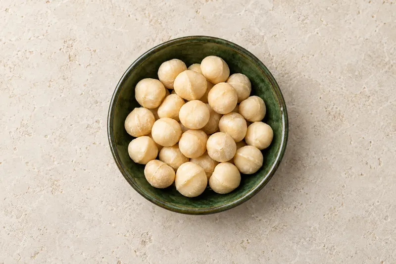 AI-generated overhead photograph-style image of ivory macadamia kernels in a shallow dark-green ceramic dish on warm pale stone.